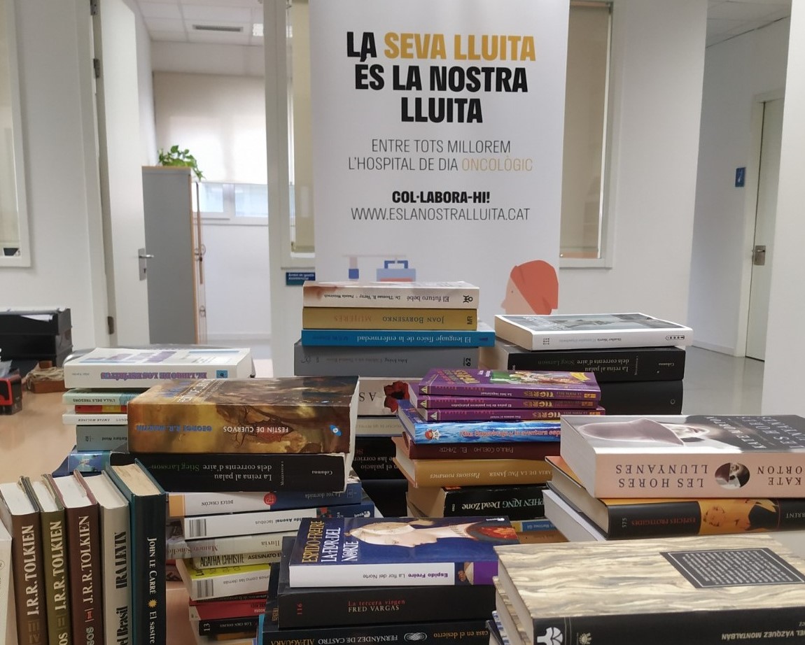 Els llibres recol·lectats apilats damunt una taula, al fons, la imatge de la campanya de mecenatge